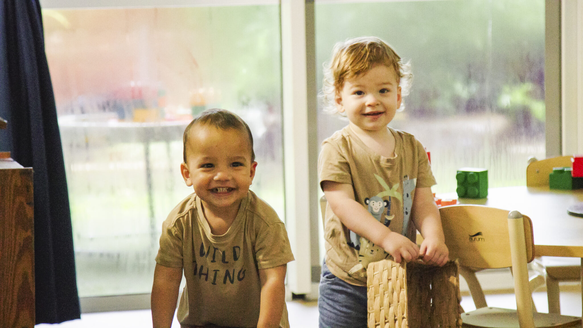 Two children standing with big smiles on their faces, inside the toddler room of the Redlynch child care centre.